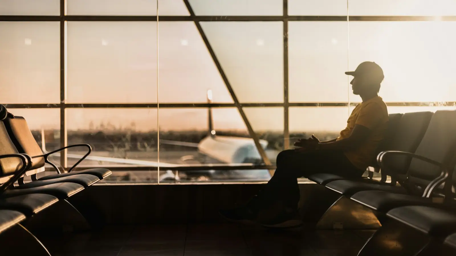 A traveler sitting alone beside a large airport window, looking out at the airfield