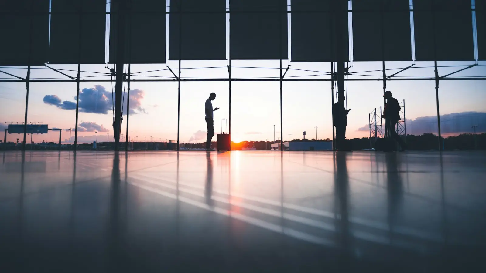 Travelers with luggage silhouetted against a wide airport terminal window at sunrise