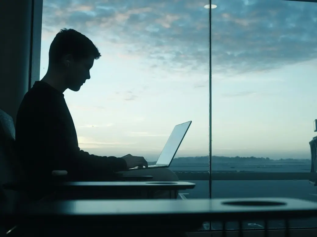A traveler working on a laptop beside an airport gate window at dawn
