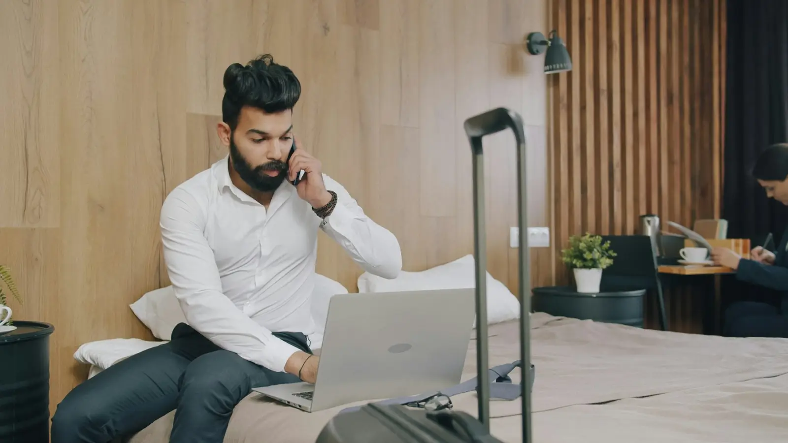A traveler with a suitcase on a phone call while checking trip details on a laptop