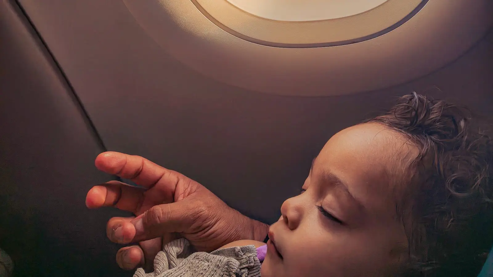 A baby asleep beside an aircraft window while an adult's hand rests close by